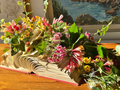 A decorative arrangement of artificial pink, yellow, and white flowers with green leaves placed on an open vintage book, set on a wooden table with a coastal painting in the background.
