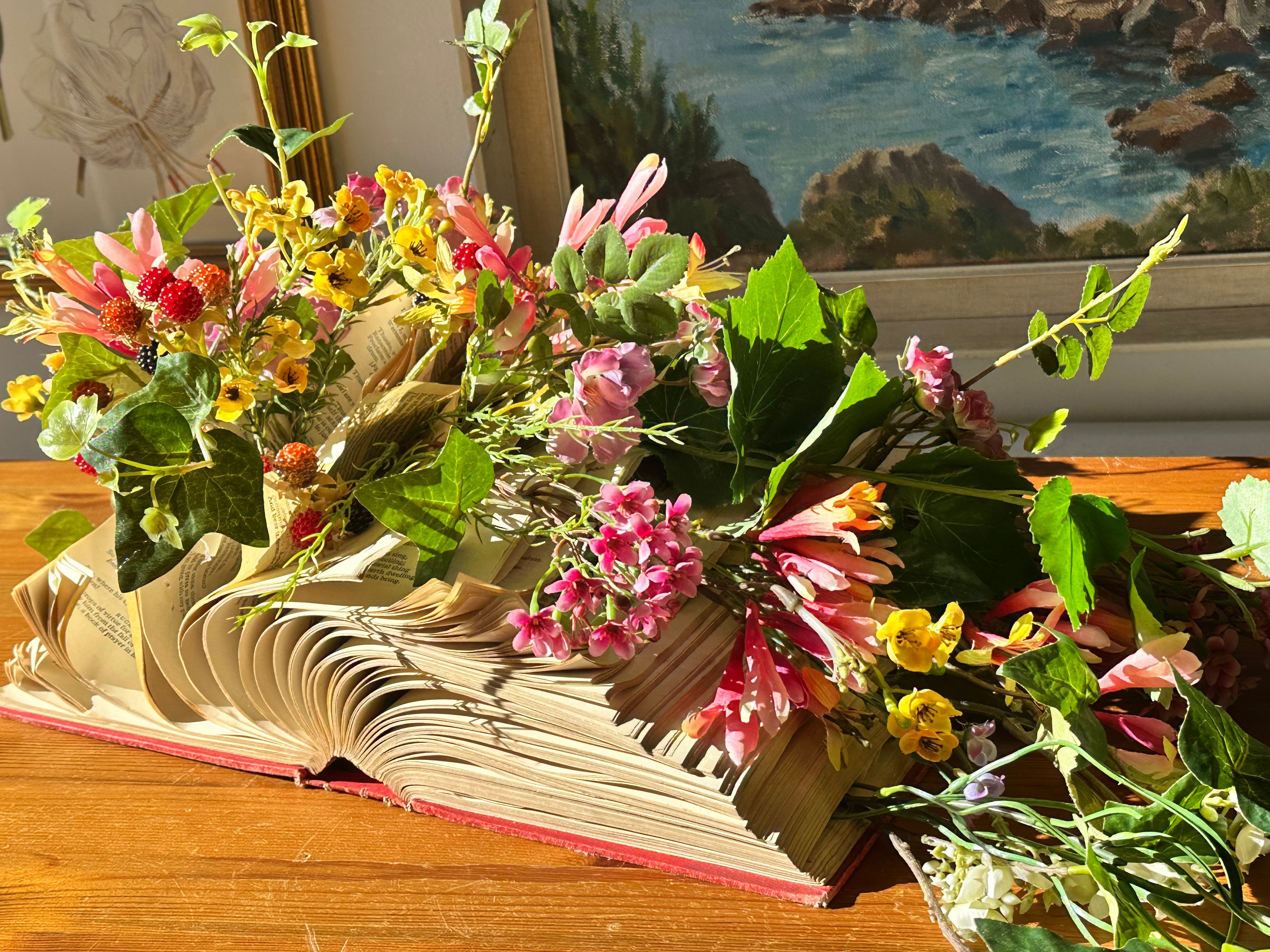 A decorative arrangement of artificial pink, yellow, and white flowers with green leaves placed on an open vintage book, set on a wooden table with a coastal painting in the background.